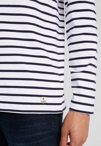 Camicia a maniche lunghe a righe bianche con righe di colore blu navy. Presenta un piccolo logo ancorato ricamato sull'orlo. Tessuto in cotone con una consistenza morbida.