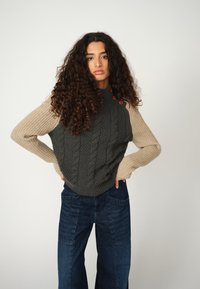 Dunkelgrauer Kabelstrickpullover mit beigen Rippenärmeln, akzentuiert durch orangefarbene Knöpfe. Kombiniert mit weit geschnittenen blauen Denim-Jeans.