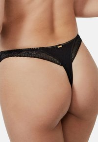Lingerie noir en string avec une bordure en dentelle et un petit accent doré. Le design présente un dos transparent avec des coutures délicates et une coupe ajustée.