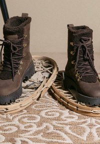 Bruine veterschoenen met een getextureerde, met bont gevoerde kraag, gouden hardware, rubberen zool en een robuust profiel, weergegeven op geweven materiaal.