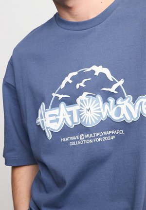 T-shirt bleu avec texte graphique blanc « HEATWAVE », des mouettes qui volent au-dessus, et un texte plus petit « HEATWAVE MULTIPLY//APPAREL COLLECTION FOR 2024. »