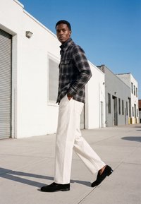 Homme marchant sur le trottoir portant une chemise à carreaux noire, un pantalon blanc et des chaussures noires près de bâtiments industriels blancs sous un ciel bleu clair.
