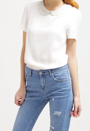 Vrouw draagt een witte blouse met korte mouwen en een met kralen versierde kraag, en lichtblauwe, hooggetailleerde, versleten spijkerbroek.