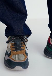 Baskets multicolores présentant un mélange de matériaux en mesh, en daim et en cuir. Le design comprend des accents gris, marron et vert, avec des superpositions texturées.