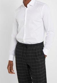 Camisa blanca de manga larga con cierre de botones combinada con pantalones gris oscuro de patrón de cuadros, con cintura elástica y cordón ajustable.