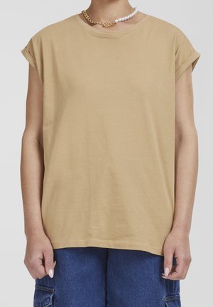 Persoon met een beige T-shirt met opgerolde mouwen, blauwe spijkerbroek en een tweekleurige ketting met gouden schakels en witte parels.