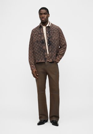 Muž stojící, oblečený v hnědém džínovém saka s vzorem, béžovém polotriku, hnědých širokých kalhotách a černých špičatých botách na bílém pozadí.