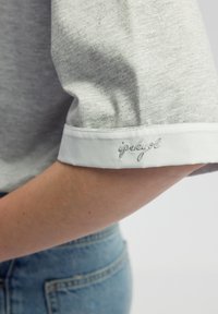 Grijs shirt met korte mouwen en een glanzende textuur, witte manchetten, en geborduurde "ipukyl" detail op de manchetten, gecombineerd met spijkerbroeken.