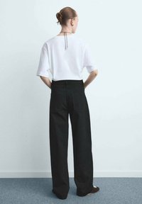 Osoba z włosami związanymi w kok, ubrana w białą koszulę z krótkim rękawem, czarne szerokie spodnie i czarne buty, stojąca na tle gładkiej jasnej ściany.