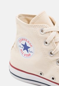 Beige canvas hoge sneakers met witte zool, rood en marineblauw streep, voorzien van een ronde blauwe logo patch met een ster en tekst.