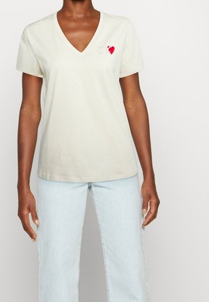 Persoon draagt een wit T-shirt met korte mouwen en V-hals met een klein rood hartje en lichtblauwe spijkerbroek tegen een effen achtergrond.
