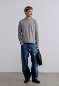 Γκρι μακρυμάνικο πουκάμισο με μικρό λογότυπο, μπλε τζιν παντελόνι με φαρδιά μπατζάκια και μαύρη τσάντα χειρός. Μονόχρωμα ρούχα, casual εφαρμογή, απαλό ύφασμα, μινιμαλιστικός σχεδιασμός.