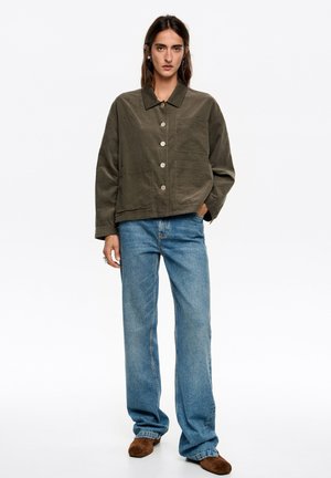 Chaqueta corta verde oliva con cuello, cinco botones grandes y dos bolsillos delanteros, combinada con jeans azules de corte recto y zapatos marrones.