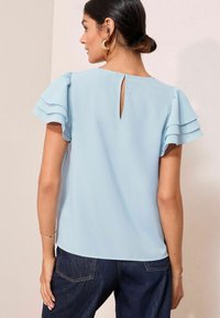 Donna con capelli scuri raccolti in un bun, che indossa una blusa azzurra chiara con volant e maniche corte, e jeans scuri, vista da dietro contro un muro semplice.