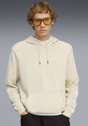 Junge Person mit lockigem Haar, die gelb getönte Brillen und einen cremefarbenen Kapuzenpullover trägt, die Hände in den Taschen, vor einem grauen Hintergrund.