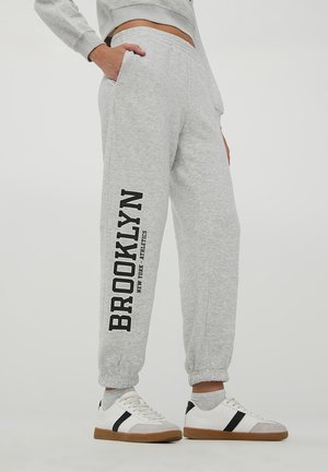 Pantaloni sportivi in cotone grigi con orli affusolati, tasche laterali e la scritta "BROOKLYN" stampata in nero sulla gamba sinistra. Indossati con sneakers bianche casual.