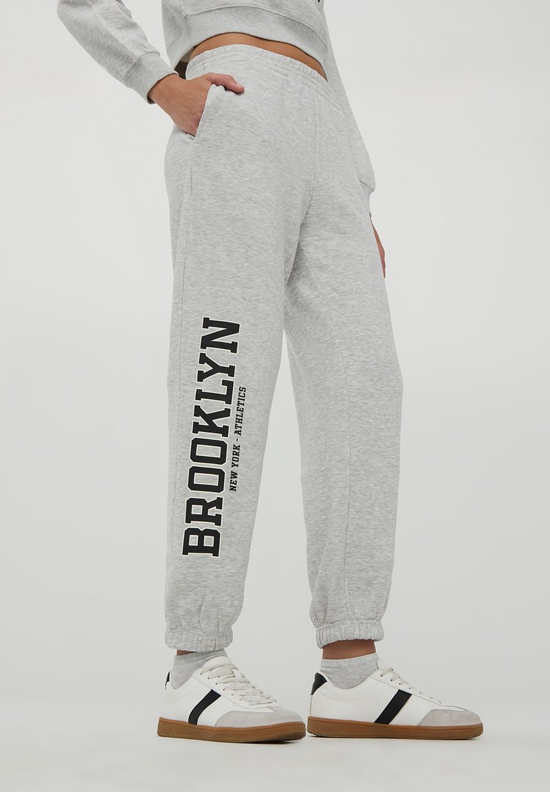 Pantaloni sportivi in cotone grigi con orli affusolati, tasche laterali e la scritta "BROOKLYN" stampata in nero sulla gamba sinistra. Indossati con sneakers bianche casual.