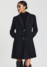 Donna che indossa un cappotto nero con bottoni e tasche, un dolcevita nero e stivali neri, in piedi contro uno sfondo chiaro e semplice.