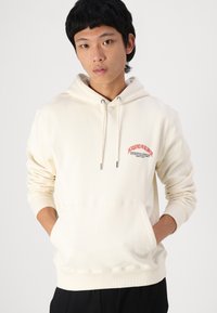 Hoodie couleur crème avec une poche kangourou à l'avant, un capuchon à cordon de serrage et un texte rouge brodé sur la poitrine. Tissu en mélange de coton doux.