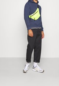 Námořnická mikina, neonově zelená kabelka přes rameno, černé zúžené kalhoty a bílé tenisky se stříbrnými a žlutými akcenty. Neformální outfit s různými texturami.