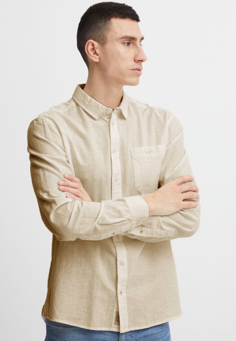 Camisa de manga larga beige, hecha de tela texturizada, con cuello tipo button-down, un bolsillo en el pecho y puños abotonados.