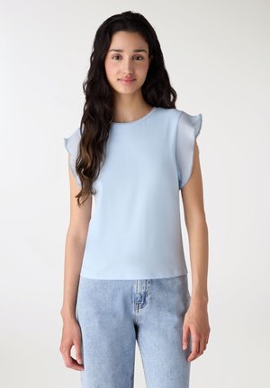 Giovane donna con lunghi capelli scuri che indossa un top azzurro senza maniche con bordi arricciati e jeans azzurri, in piedi davanti a uno sfondo semplice.