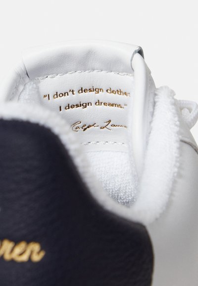 Biały skórzany sneaker z granatowymi akcentami, z fakturowanym językiem i złotym napisem wewnątrz: "Nie projektuję ubrań; projektuję marzenia."