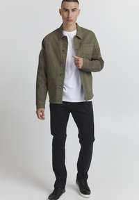 Olivnozeleni denim jakna z gumbom spredaj, prsno žepom in dvema stranskima žepoma, nosi se preko bele majice in v kombinaciji z črnimi hlačami.