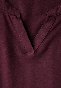 Haut bordeaux avec décolleté en V, tissu doux texturé et couture discrète autour du col. Vue en gros plan.