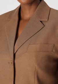 Brun strukturert blazer med notched lapel, enkelt knappelukking, og brystlomme. Stoffet har en glatt, skreddersydd tekstur.