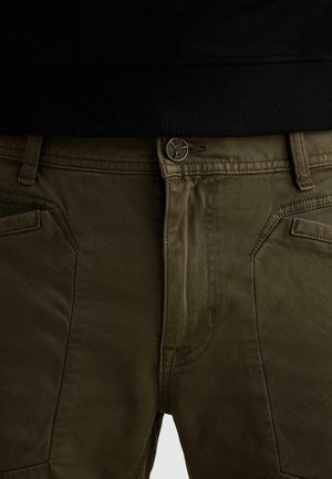 Gros plan sur un pantalon vert olive avec une couture détaillée, une fermeture à boutons en métal et des poches avant sous un vêtement noir.