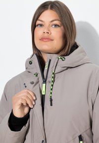 Donna che indossa una giacca leggera grigia con cappuccio e lacci di un verde neon, mentre tiene il colletto della giacca contro uno sfondo bianco.