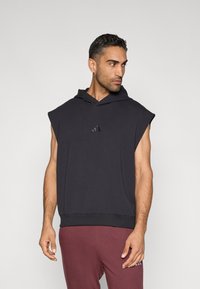 Juodas be rankovių džemperis su kapišonu ir ribuotu apvadu, ant krūtinės mažytis juodas Adidas logotipas. Lygus audinys, minimalistinis dizainas.