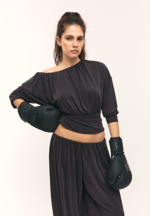 Žena s čiernymi boxerskými rukavicami a voľným čiernym outfitom s odhalenými ramenami, stojaca s jednou rukou na bokoch pred jednofarebným pozadím.