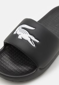 Infradito neri con una soletta testurizzata, parte superiore in sintetico liscio e un logo in bianco a forma di coccodrillo sulla cinghia.