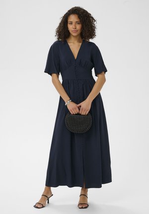 Femme aux cheveux bouclés portant une robe midi bleu marine boutonnée et des sandales à talons noires, tenant un sac à main tissé noir devant elle.