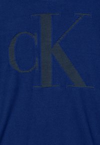 Plava tkanina s istaknutim, izvezenim tamnosivim logotipom "CK" djelomično centriranim, s glatkom teksturom i dosljednim uzorkom tkanja.
