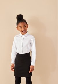 Chemise blanche à manches longues avec boutons, associée à une jupe noire. La chemise présente un col et la jupe possède une texture lisse.