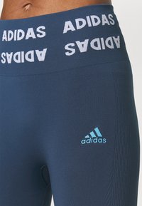 Leggings atléticos de color azul marino con una cintura ancha que presenta el texto "ADIDAS" en blanco y un logo en azul claro en la parte inferior de la pierna. Tejido suave y elástico.