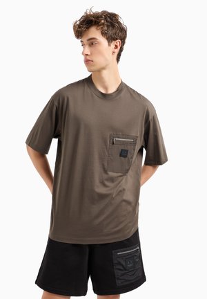 T-shirt marrone a maniche corte realizzato in tessuto morbido, con tasca con zip sul petto sinistro e vestibilità comoda. Abbinato a pantaloni corti neri.
