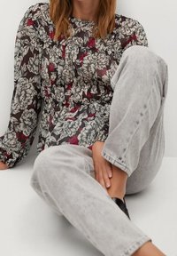 Blusa nera trasparente con motivo floreale bianco e rosso, scollo rotondo, maniche lunghe, abbinata a jeans grigi chiari e scarpe slip-on nere.