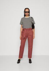 Schwarz-weißer gestreifter T-Shirt mit kurzen Ärmeln, kombiniert mit einer hoch taillierten Hose in Rostfarbe mit einem Bindegürtel in der Taille. Schwarze Sonnenbrille und Tasche.