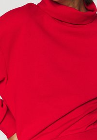 Czerwony bawełniany top z wysokim, podwiniętym dekoltem i luźnymi rękawami. Tkanina jest miękka o gładkiej fakturze, a krój jest luźny.