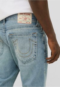 Jeans de mezclilla azul claros con un corte relajado, que cuentan con costuras detalladas y un parche con el logo en la parte trasera de la cintura. Se pueden ver dos bolsillos traseros.