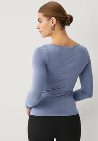 Camiseta de manga larga en suave tejido azul claro, con escote redondo y detalles fruncidos en los laterales. Combinada con leggings negros.