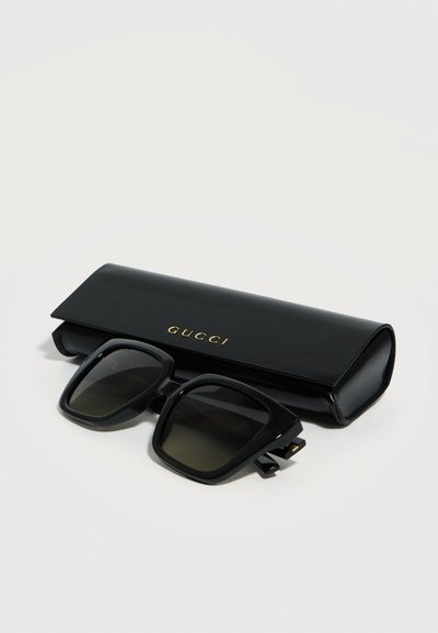 Μαύρα τετράγωνα υπερμεγέθη γυαλιά ηλίου με σκοτεινούς φακούς που ακουμπούν μπροστά σε σκληρή θήκη Gucci σε μαύρο χρώμα με χρυσά γράμματα.