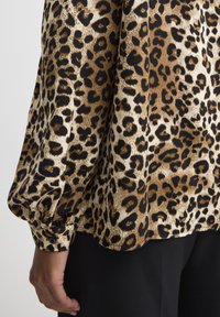 Blouse à imprimé léopard mêlant des tons marron, crème et noir. Manches longues bouffantes avec détail de poignet bouton. Tissu doux et texturé.