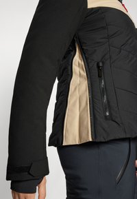 Svart isolert jakke med quilted design, beige innvendig, glidelåsdetaljer og borrelås-lukking på ermene.