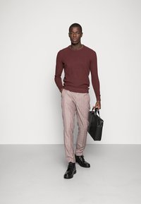 Bordeauxfarbener Pullover, lichte kariert Hose und schwarze Stiefel. Trägt eine schwarze Aktentasche. Insgesamt ein smart-casual Outfit mit modernem Schnitt.