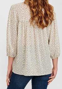 Blouse florale légère présentant un fond crème avec des motifs violets et verts, des manches trois-quarts et une coupe ample et froncée.